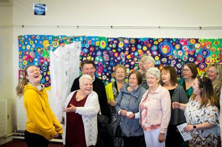 A group of older participants with two of the artists, standing in front of a large painting at the Roam exhibition in Mayfield Library