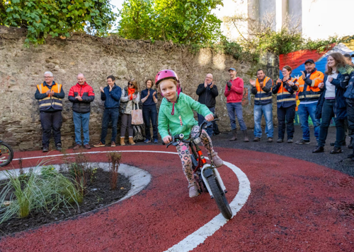 A young girl cycles in a external space, Railway Park, while local residents and Cork City Council staff cheer her on.