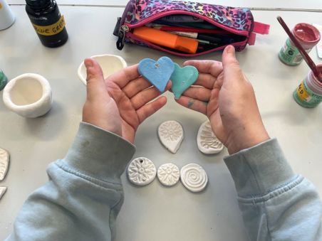 A child's hands holding two painted clay hearts, with several unpainted clay tiles on a table.